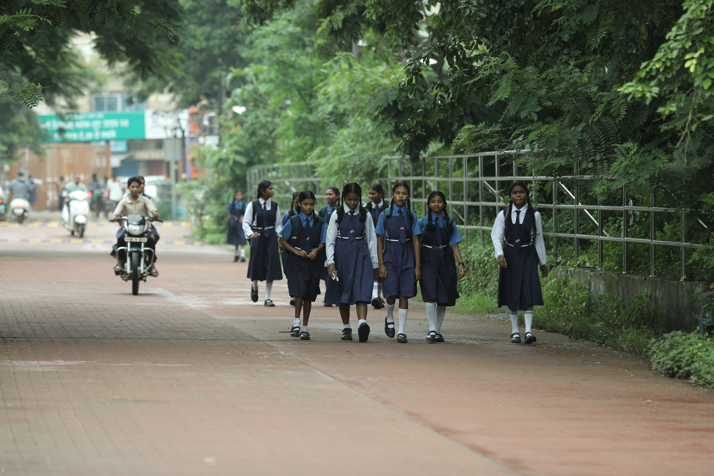 Schoolgirls in navy uniforms walking together along a quiet, tree-lined path on their way to school.